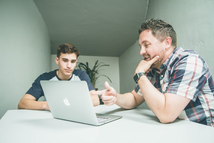 Two men seated at a laptop with one explaining something to the other